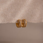 Ribbed Pave Huggies featuring a textured gold finish and embedded cubic zirconia stones, shown on a soft beige surface—ideal earrings for those who love statement jewelry.