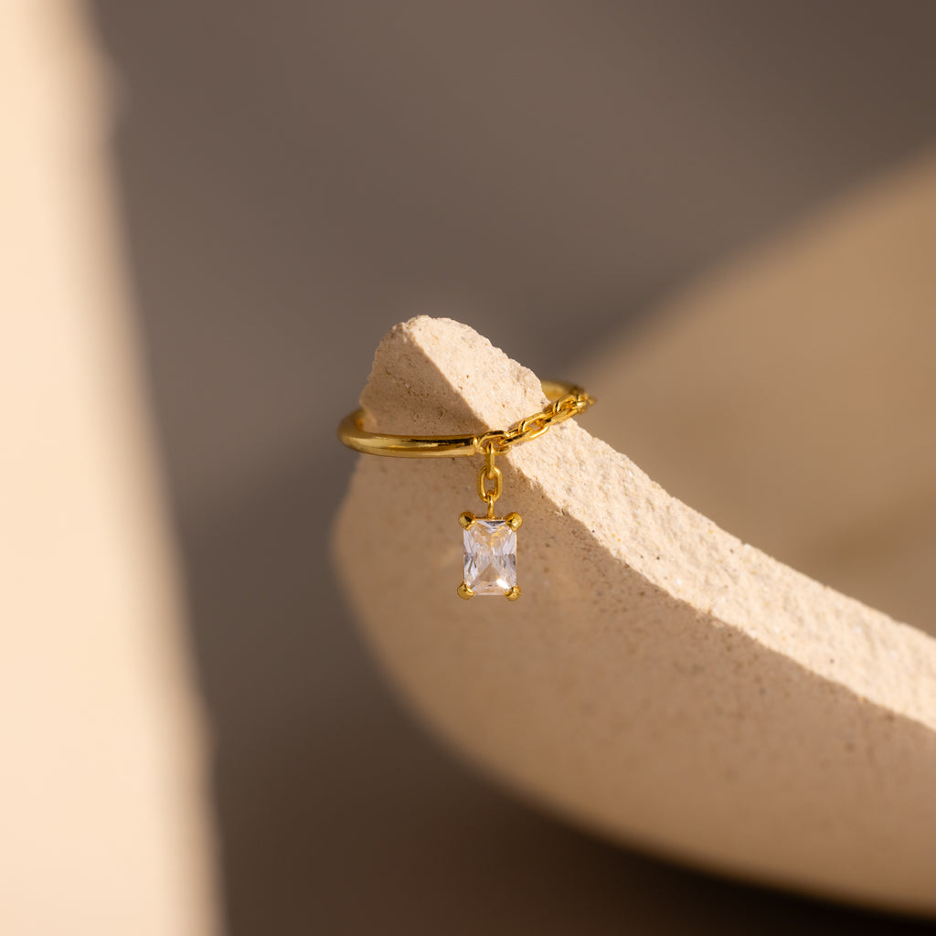 Diamond Drop Chain Ring with a dangling rectangular emerald-cut stone, displayed on a textured beige surface in soft light.