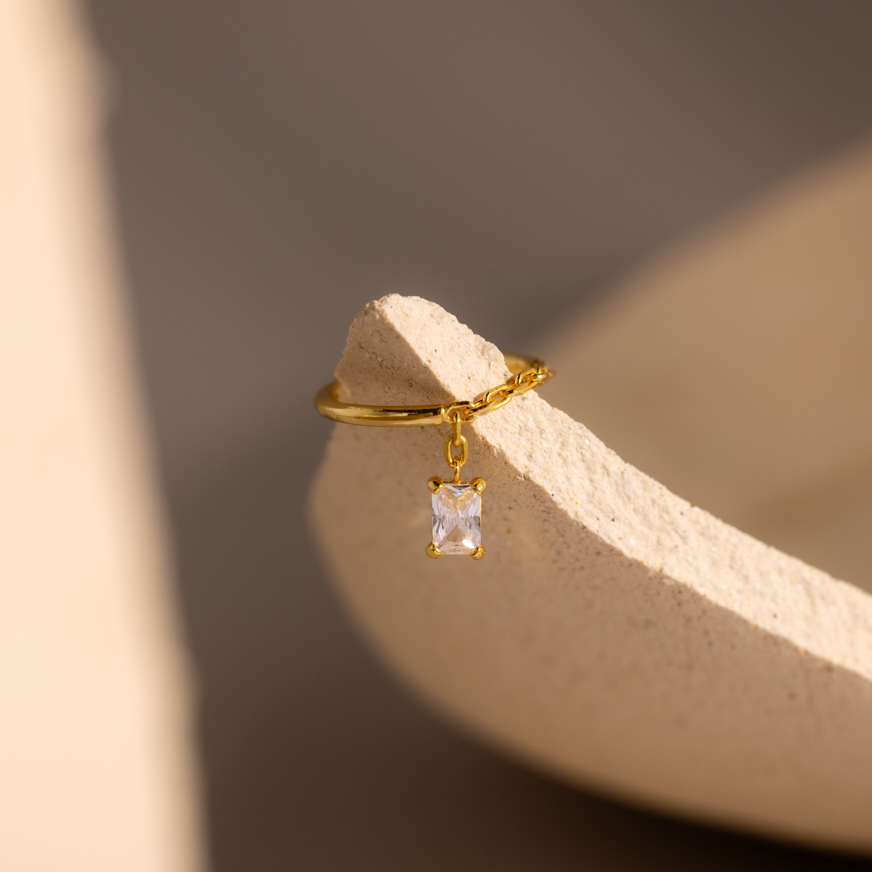 Diamond Drop Chain Ring with a dangling rectangular emerald-cut stone, displayed on a textured beige surface in soft light.