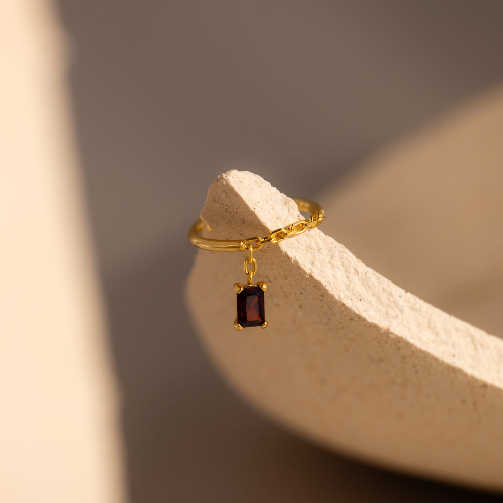 The Garnet Drop Chain Ring features a garnet stone pendant, elegantly resting on the edge of a textured beige surface.