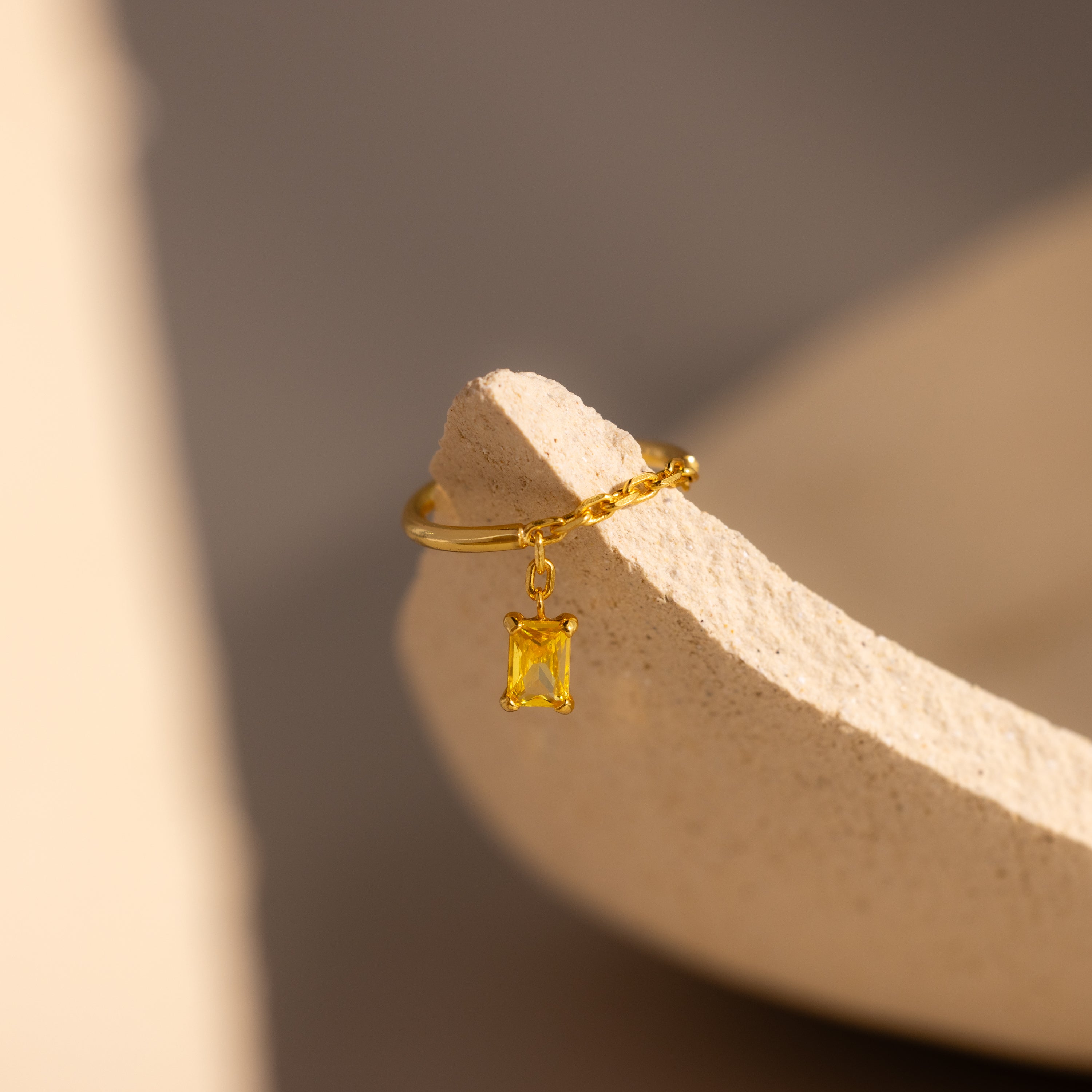 Topaz Drop Chain Ring in gold, featuring an emerald-cut stone charm, beautifully displayed on a textured beige ceramic surface.
