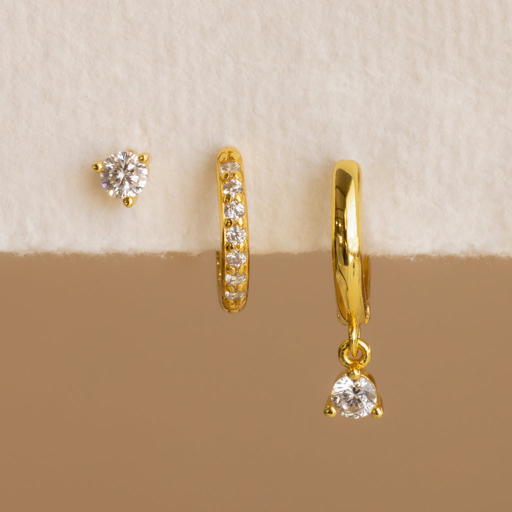 The Classic Diamond Earrings Set, featuring three mismatched gold earrings—including a classic stud with clear gemstones—is displayed on a white and beige textured surface.