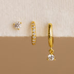 The Classic Diamond Earrings Set, featuring three mismatched gold earrings—including a classic stud with clear gemstones—is displayed on a white and beige textured surface.