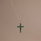 Silver chain necklace featuring a cross pendant set with small, round green stones against a neutral background.