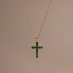 Gold chain necklace featuring a cross pendant set with small, round green stones against a neutral background.