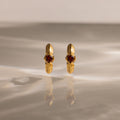 The Ribbed Garnet Huggies, gold hoop earrings with red gemstones, stand upright on a softly lit reflective surface—making them a perfect meaningful gift.