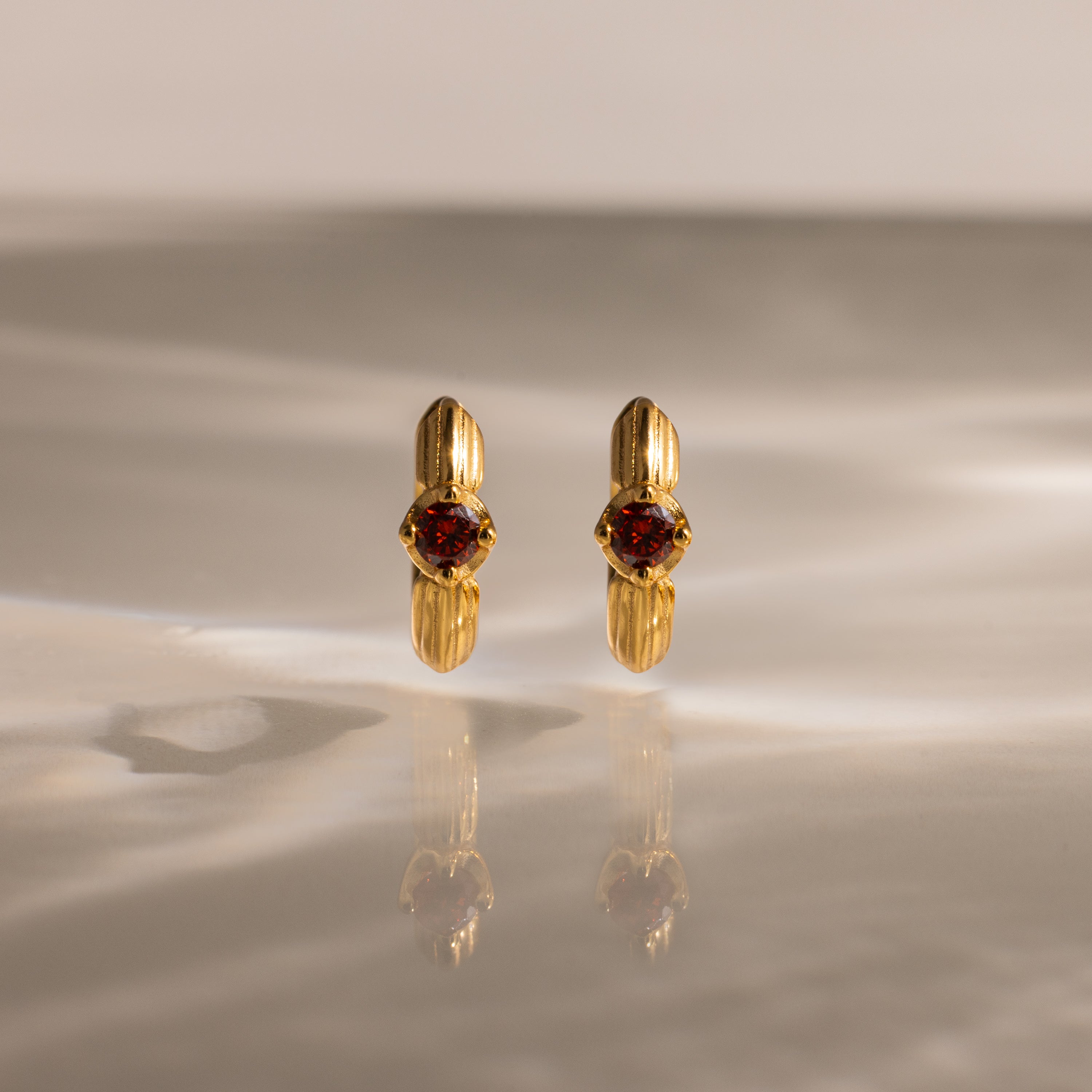 The Ribbed Garnet Huggies, gold hoop earrings with red gemstones, stand upright on a softly lit reflective surface—making them a perfect meaningful gift.