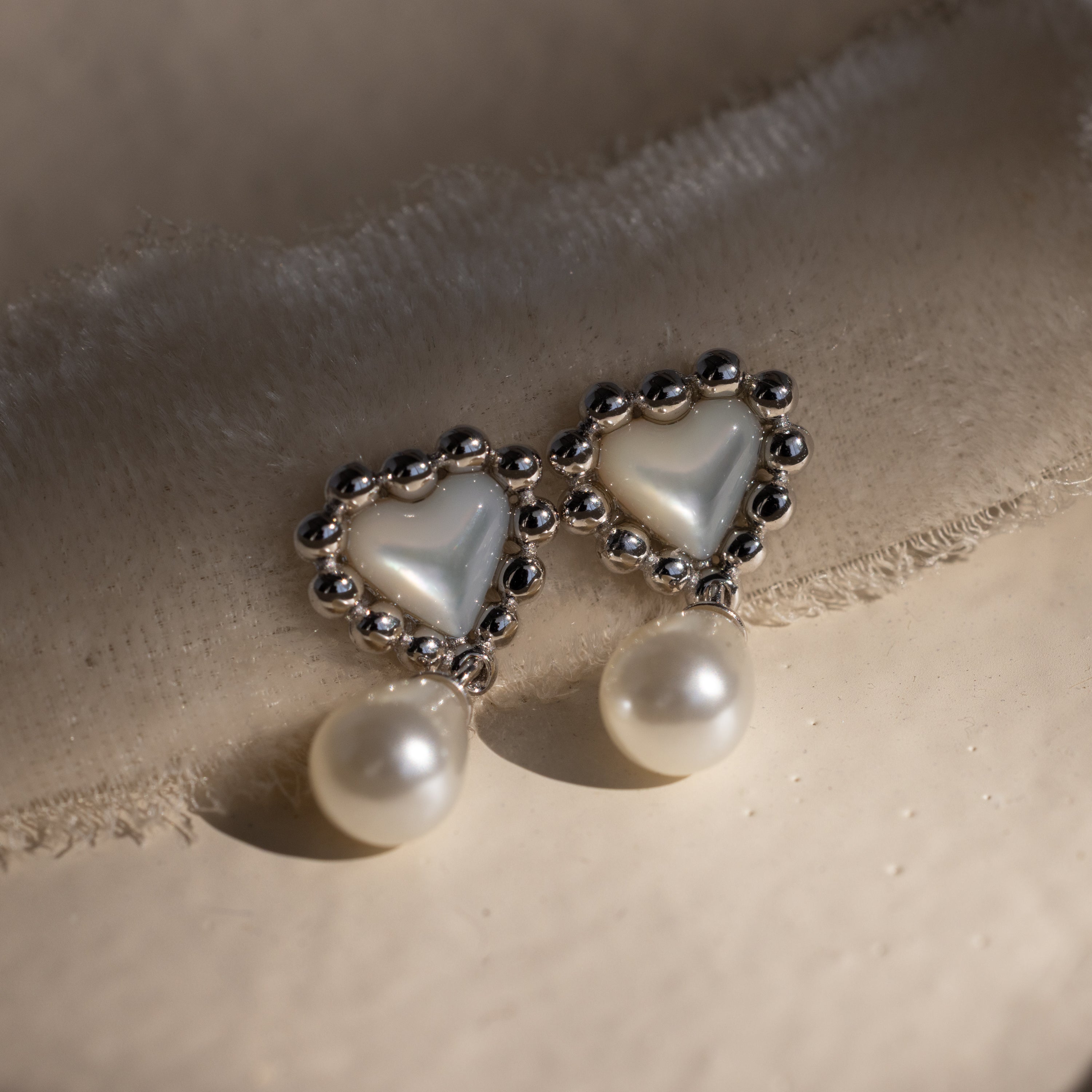 Flat lay of silver beaded heart earrings with pearl drops, arranged in warm sunlight on a beige fabric surface.