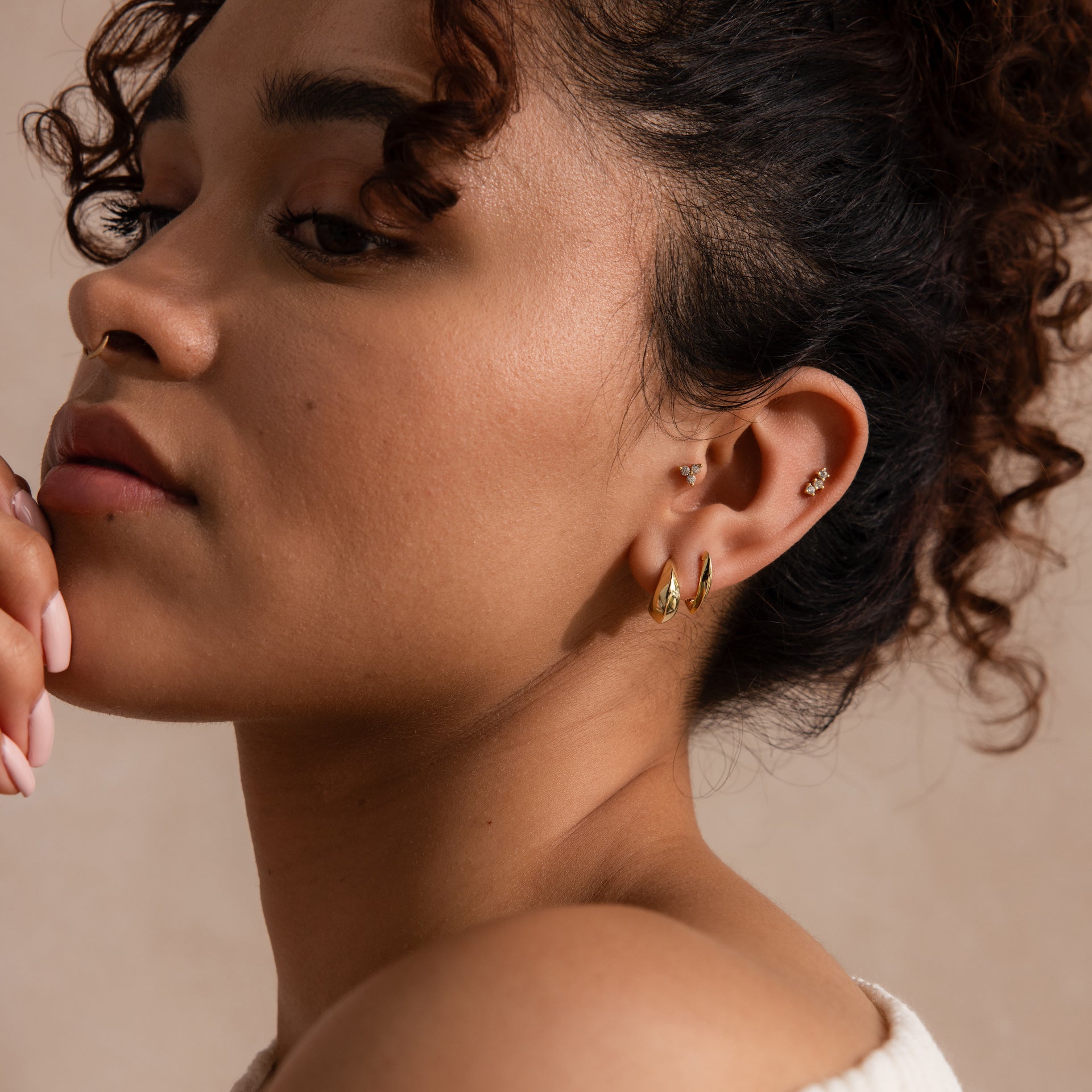 Model wearing stacked gold earrings, including sculptural oval-shaped huggies and delicate pavé studs.