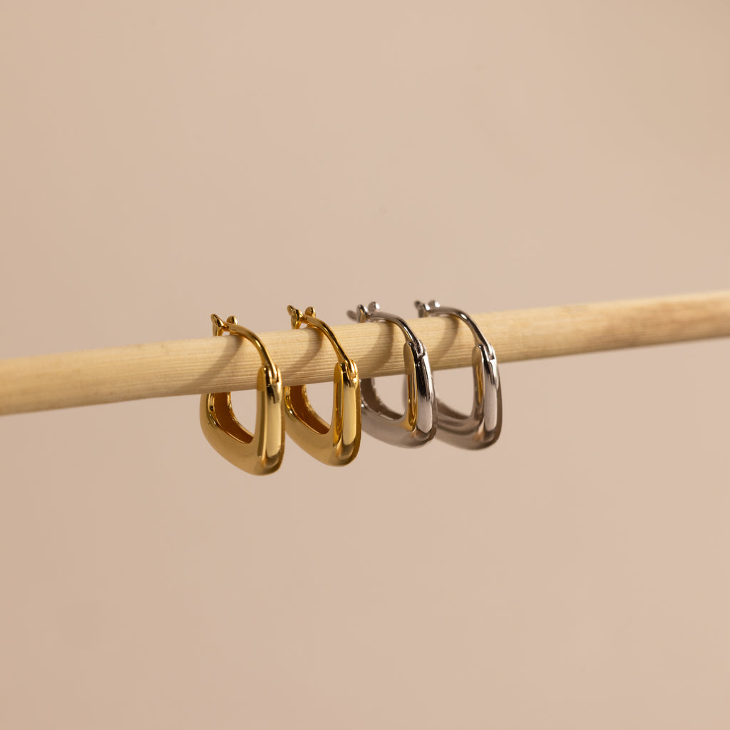 Gold and silver geometric square hoop earrings displayed side by side on a wooden rod, showcasing their bold modern shape and sleek finish.