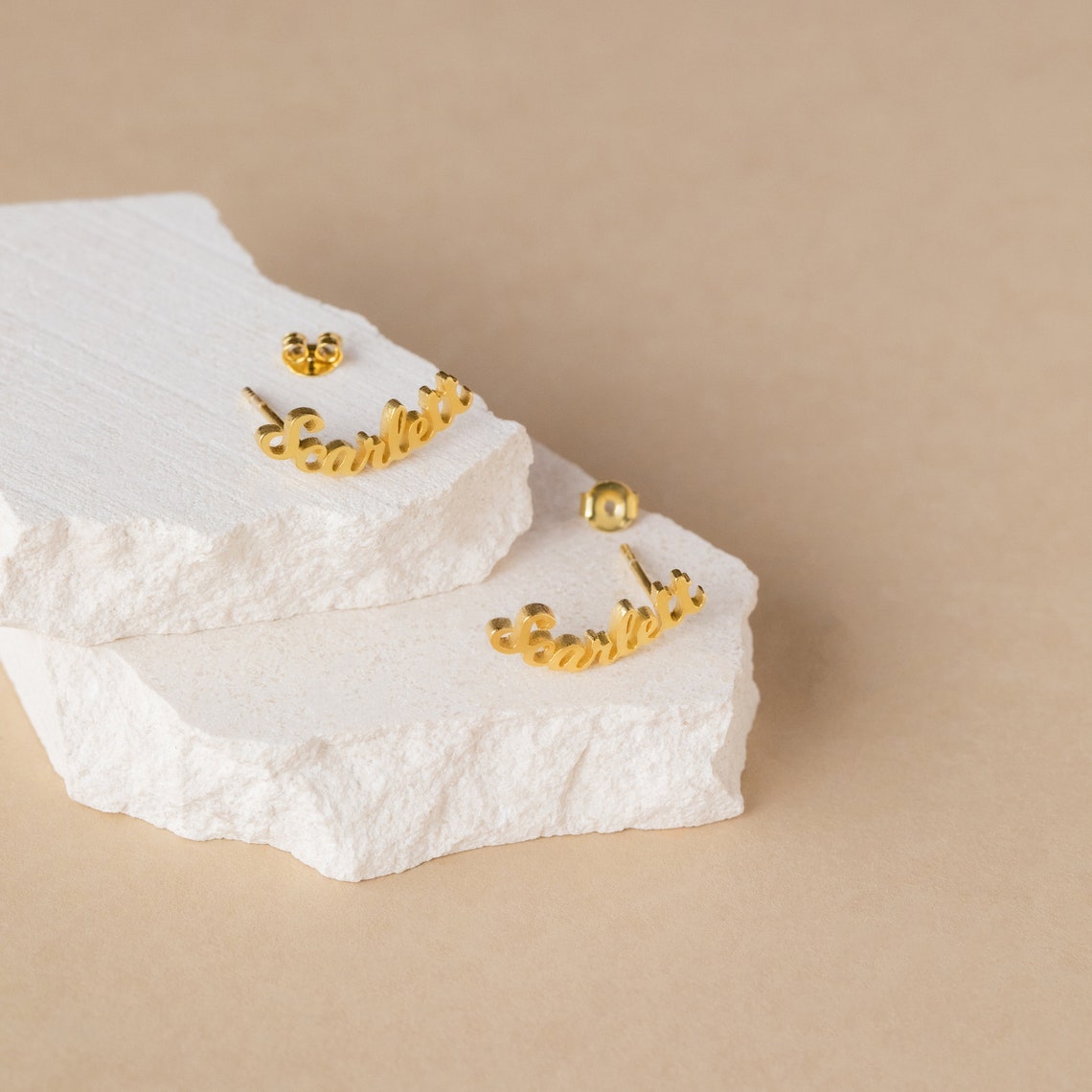 Gold cursive name earrings on a textured white surface with a beige background