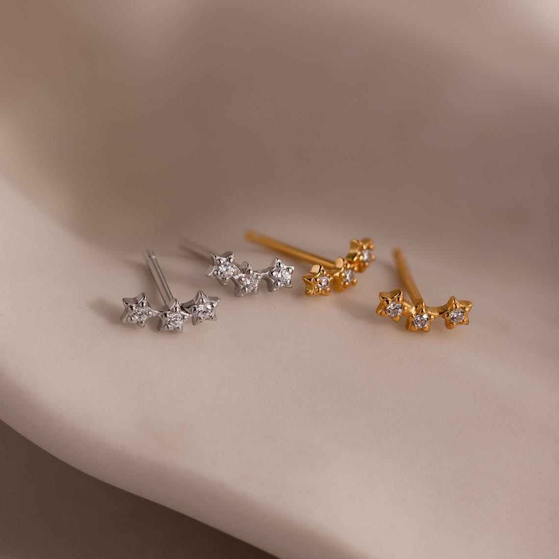 Two pairs of star-shaped earrings in gold and silver on a neutral background