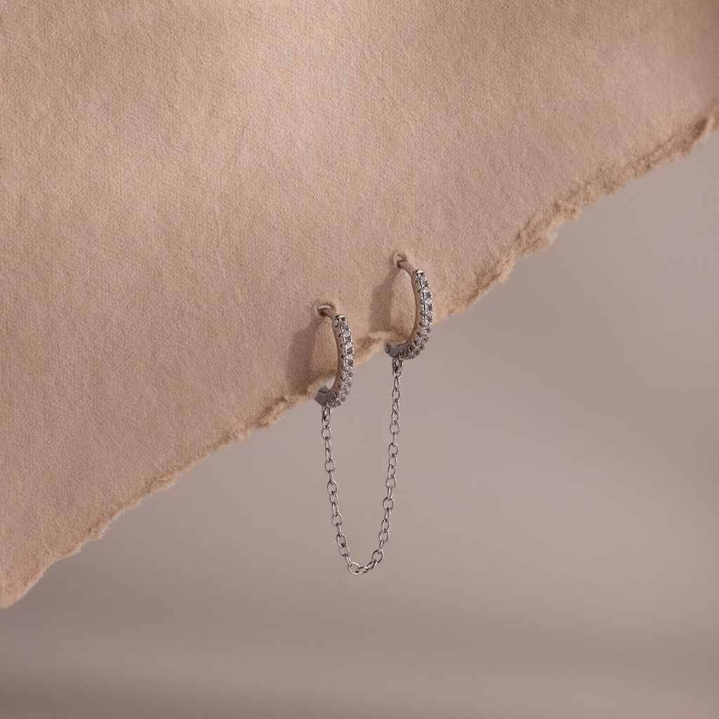 Silver pave hoop earrings with chain detail on a textured beige surface