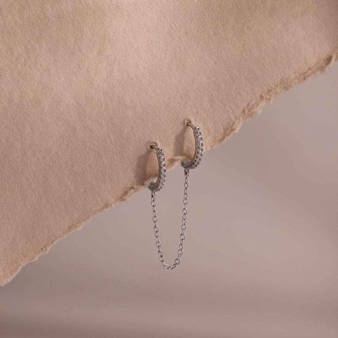 Silver pave hoop earrings with chain detail on a textured beige surface