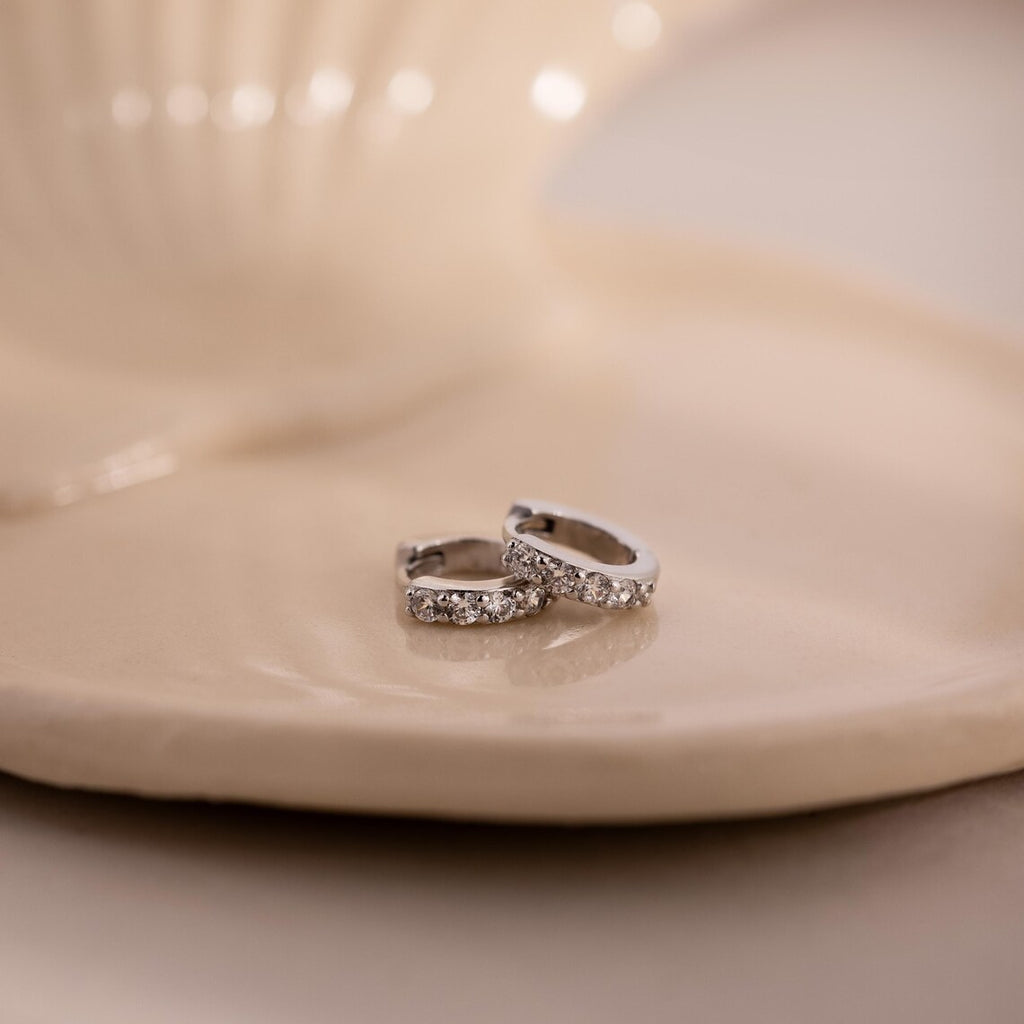 Two silver earrings with clear gemstones on a beige ceramic plate.