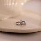 Two silver earrings with clear gemstones on a beige ceramic plate.