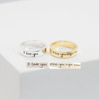 A silver Handwriting Band engraved with "I love you" and a gold custom ring with "miss you right now" rest on a white surface—ideal as heartfelt Christmas gifts.