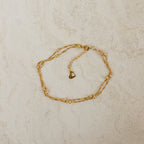 The Duo Bead Chain Bracelet, featuring a delicate gold chain with small beads and a heart-shaped clasp, rests on light marble—perfect to pair with your favorite necklace for a chic, layered look.