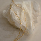 The Duo Bead Chain Anklet features delicate gold beading, elegantly draped over a white crystal stone on a neutral background, showcasing its effortless and dainty design.