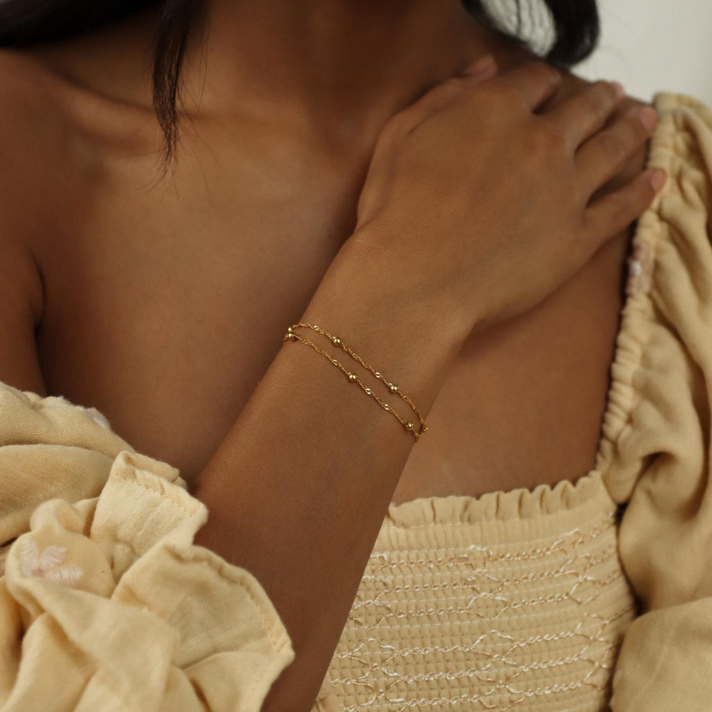 A woman wears the Duo Bead Chain Bracelet with a cream off-shoulder, textured dress featuring puffed sleeves.