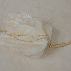 The Duo Bead Chain Bracelet pairs beautifully with a delicate gold chain necklace draped over a white crystal stone on a beige surface, creating an effortlessly chic and elegant look.