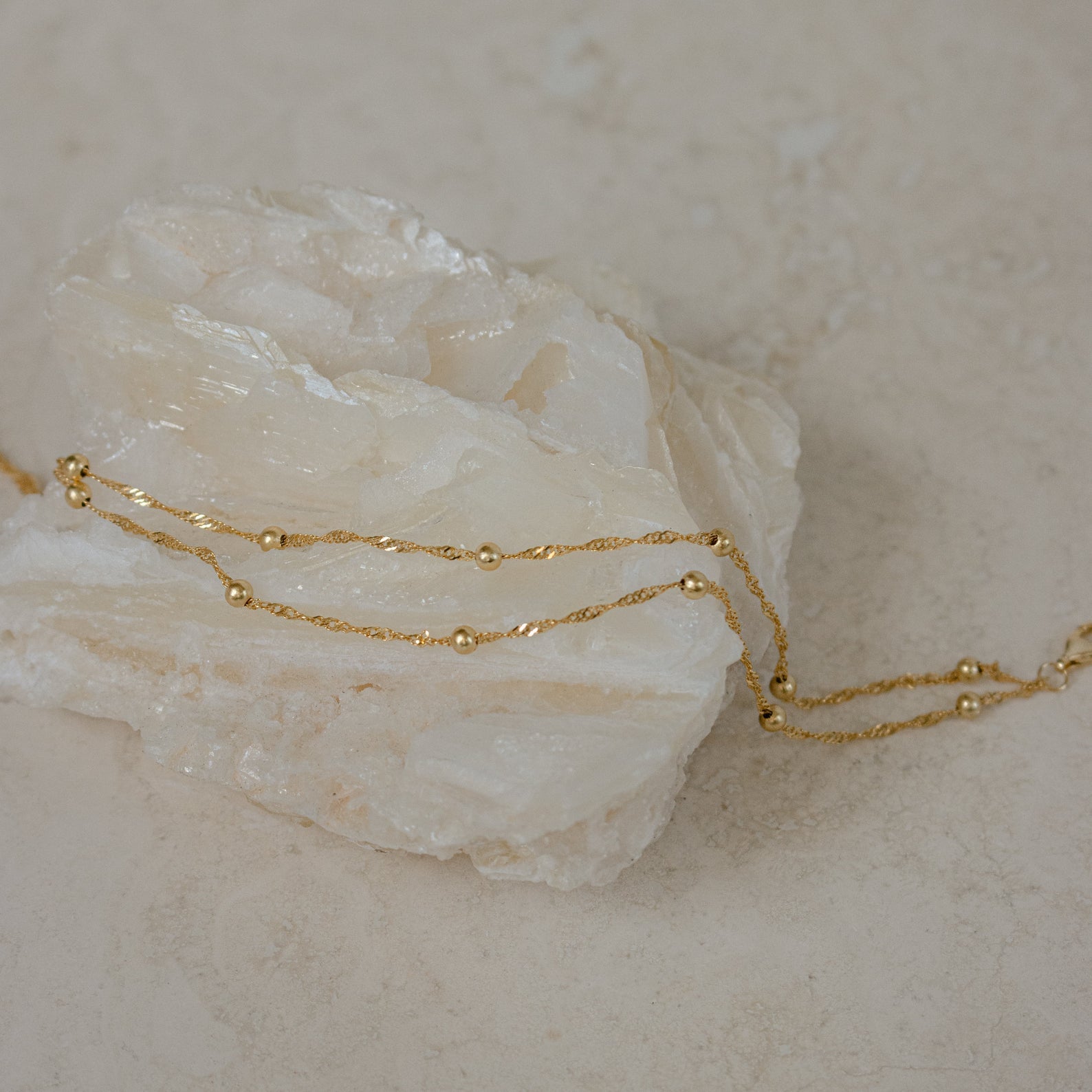 The Duo Bead Chain Bracelet pairs beautifully with a delicate gold chain necklace draped over a white crystal stone on a beige surface, creating an effortlessly chic and elegant look.