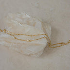 The Duo Bead Chain Anklet in Sterling Silver features delicate small beads draped elegantly over a rough white crystal on a neutral background.