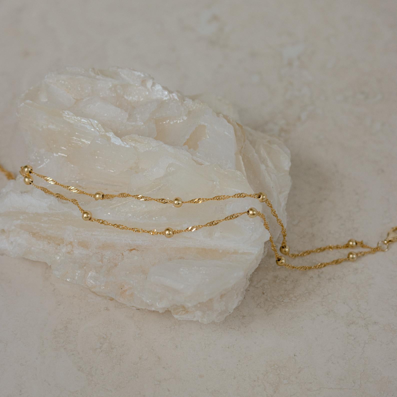 The Duo Bead Chain Anklet in Sterling Silver features delicate small beads draped elegantly over a rough white crystal on a neutral background.