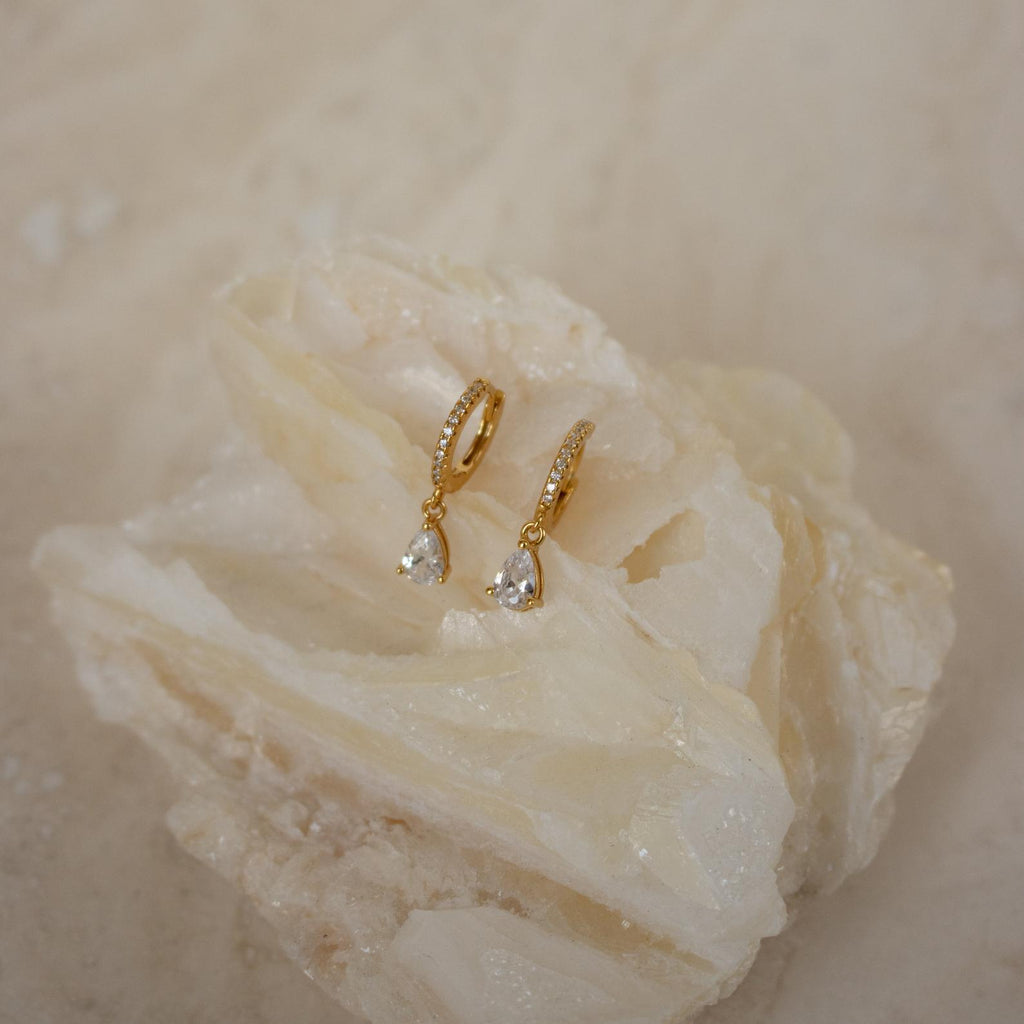 Diamond Drop Huggies in 18K Gold with teardrop gemstones shimmer on a textured, light surface—ideal for bringing elegance to semi-formal outfits.