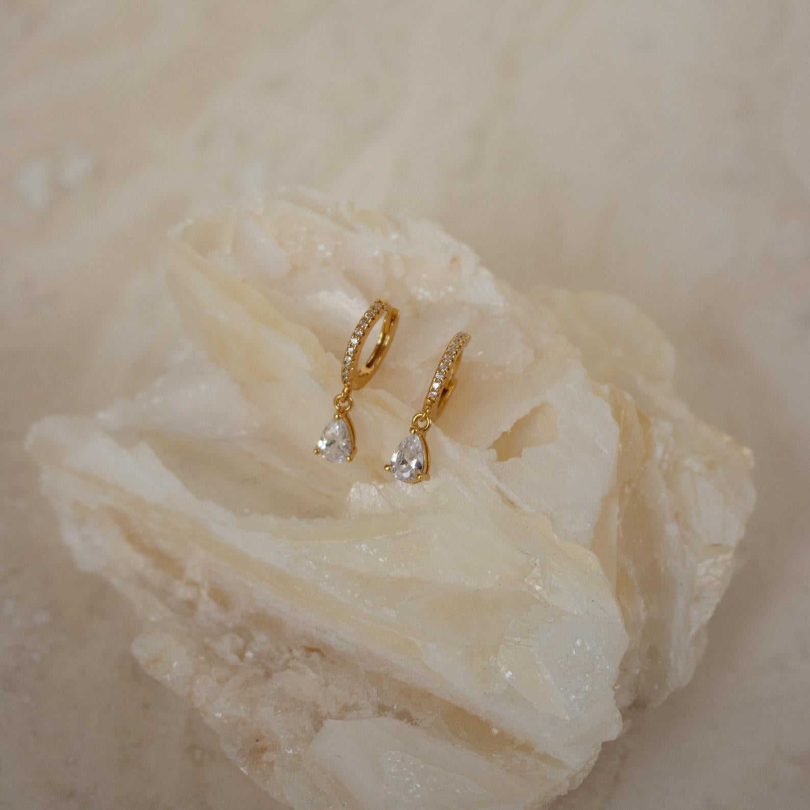Diamond Drop Huggies in 18K Gold with teardrop gemstones shimmer on a textured, light surface—ideal for bringing elegance to semi-formal outfits.