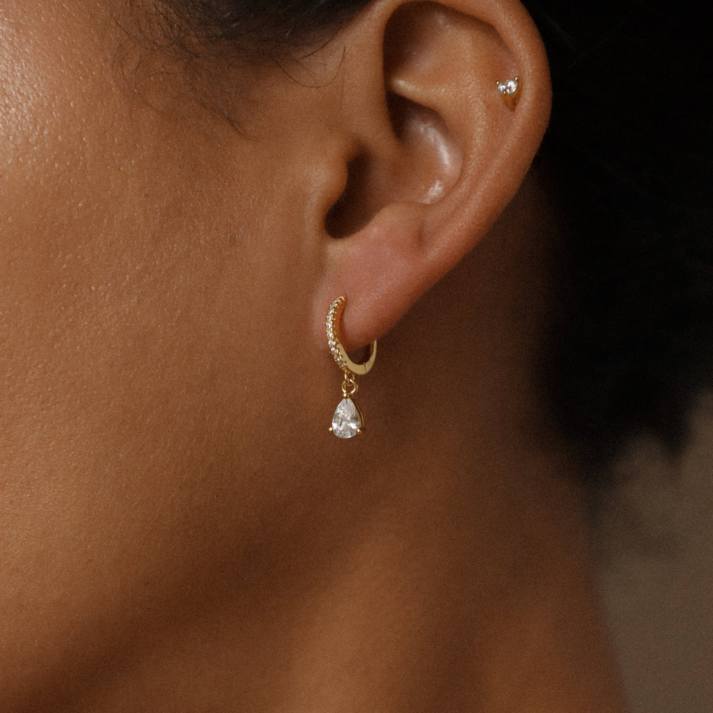 Close-up of a person’s ear wearing Diamond Drop Huggies in 18K Gold featuring a teardrop-shaped pendant and tiny stud—an elegant choice for semi-formal attire.