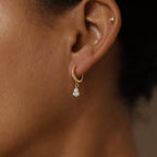 Close-up of a person’s ear wearing Diamond Drop Huggies in 18K Gold featuring a teardrop-shaped pendant and tiny stud—an elegant choice for semi-formal attire.