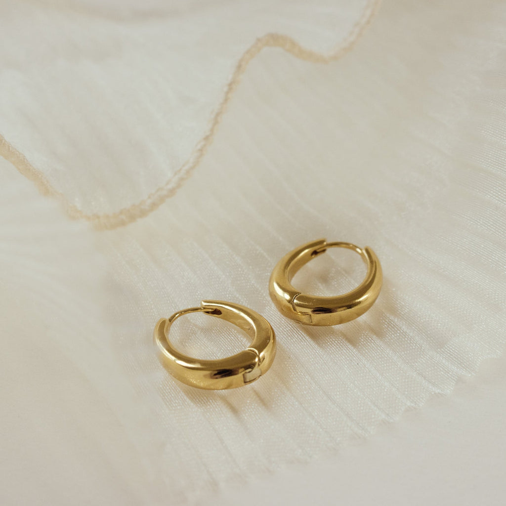 Paris Oval Hoops in 18K Gold shown on a textured cream backdrop—these classic earrings bring timeless elegance to your style.