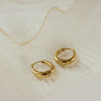 The Paris Oval Hoops, classic gold earrings, are displayed elegantly on a white, pleated fabric background.