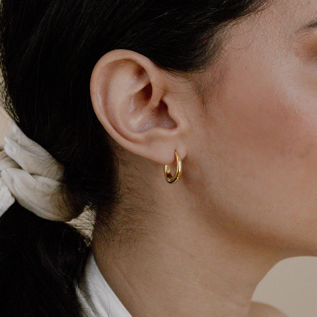 Close-up of a woman's ear wearing Paris Oval Hoops, with dark hair tied back in a light scarf.