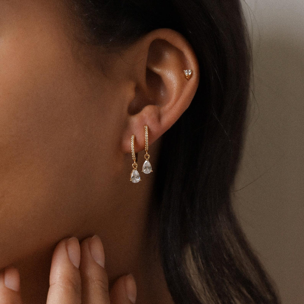 Close-up of a woman's ear wearing Diamond Drop Huggies in 18K Gold—diamond-encrusted hoops with clear gemstone drops—paired with an upper ear stud, ideal for semi-formal looks.