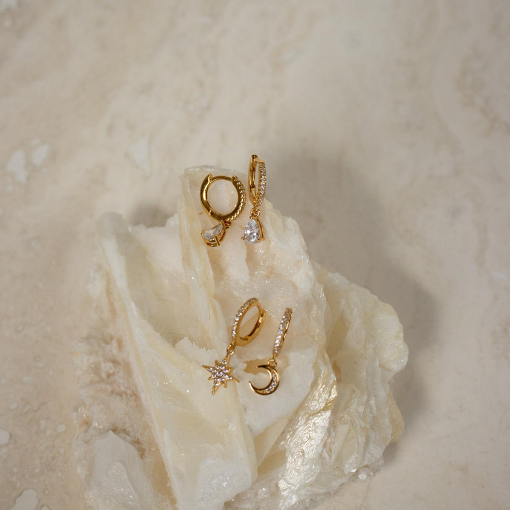The Diamond Drop Huggies in 18K Gold, along with two other elegant gold earring pairs, are showcased on textured white stone—an ideal accent for semi-formal outfits.