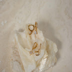The Diamond Drop Huggies in 18K Gold, along with two other elegant gold earring pairs, are showcased on textured white stone—an ideal accent for semi-formal outfits.