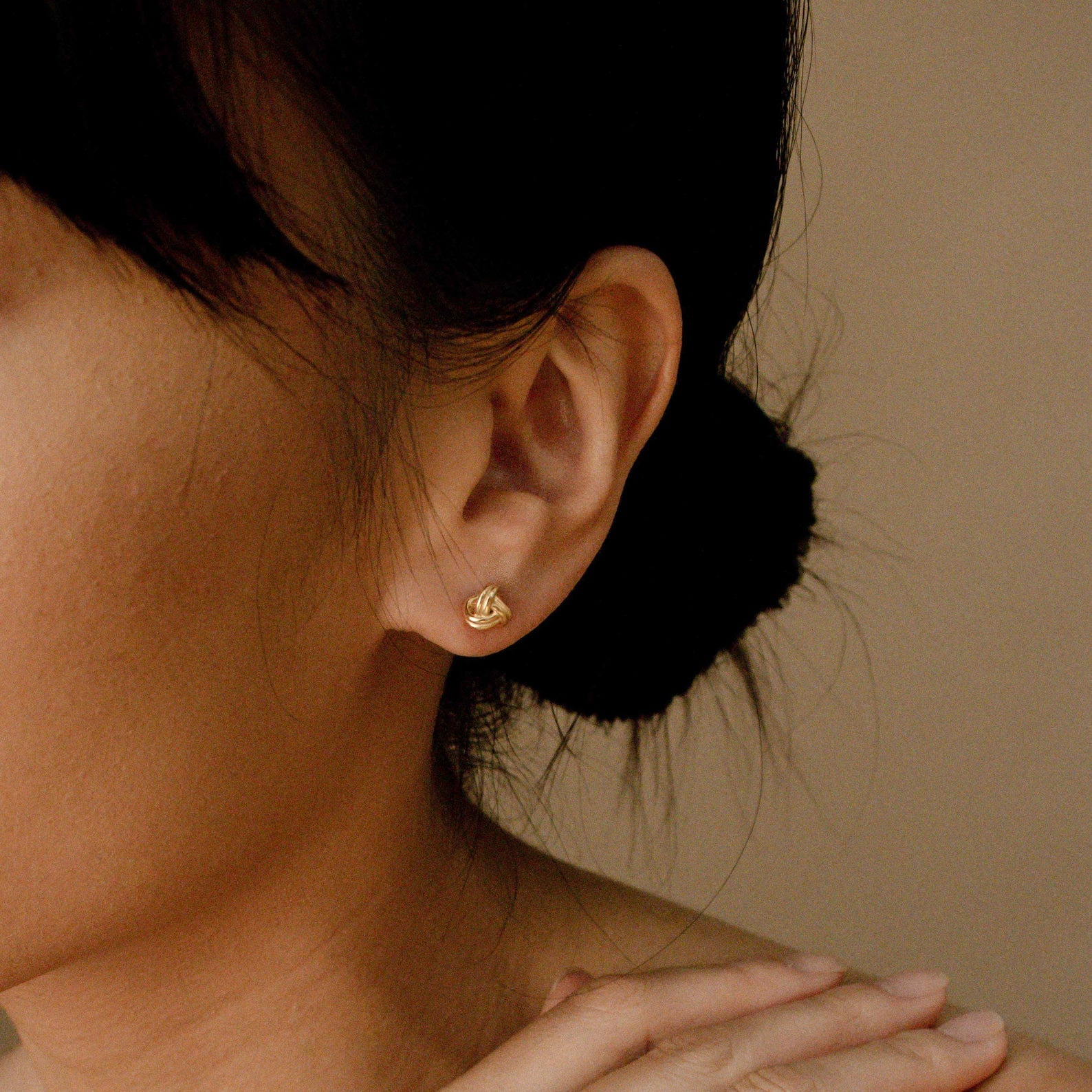 Close-up of a woman with dark hair in a bun wearing Love Knot Studs in 18K Gold—symbolic earrings that make a thoughtful gift—her hand resting gently near her neck.