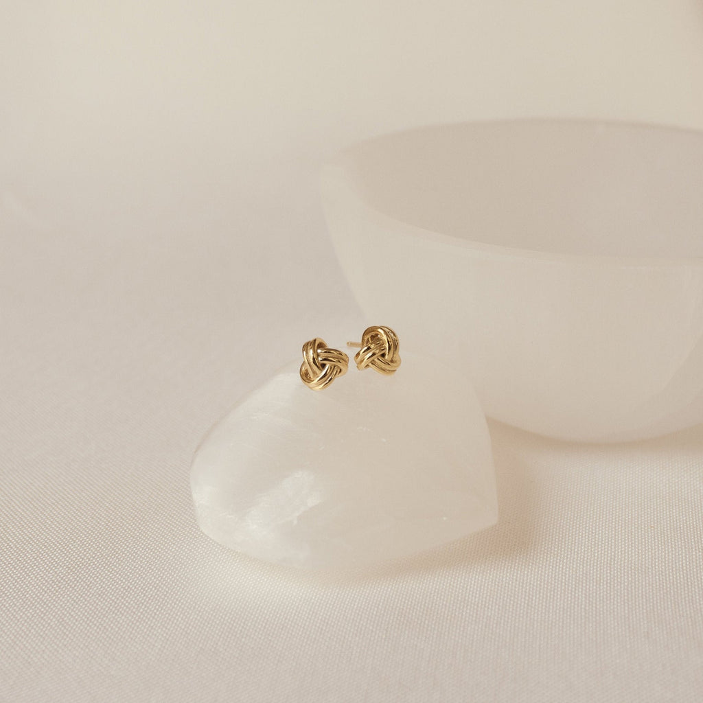 Love Knot Studs in 18K Gold displayed on a smooth white stone with a round white bowl in the background—ideal as a meaningful gift or cherished symbolic jewelry.