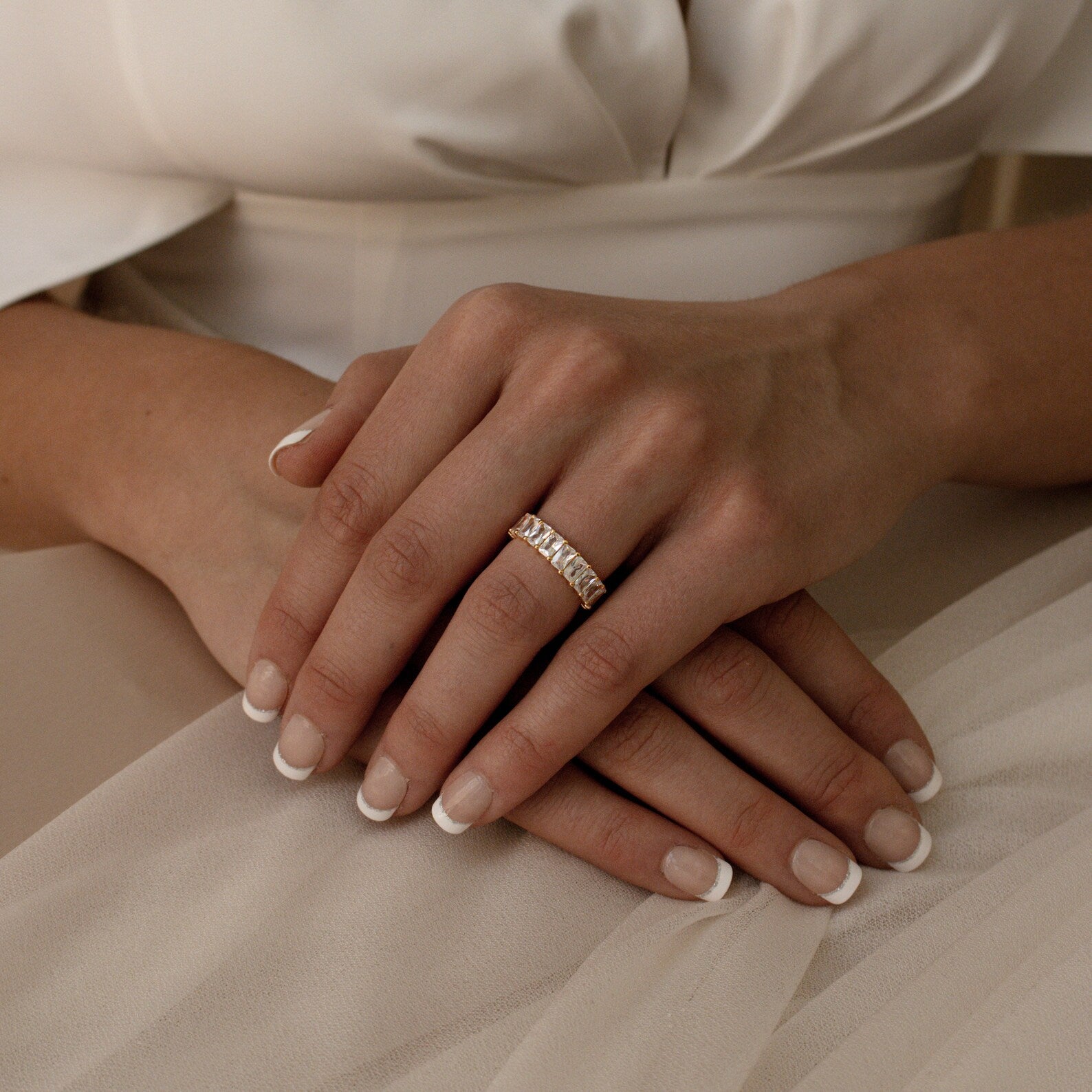 Close-up of hands with French manicured nails wearing the Colette Eternity Ring, an opulent gold band set with baguette diamonds, resting on a white dress.