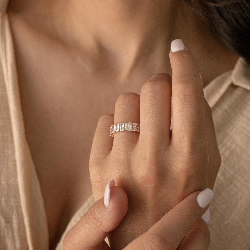 A hand with white nail polish wears the Colette Eternity Ring, a gold band adorned with multiple rectangular baguette diamonds for a look of subtle opulence.