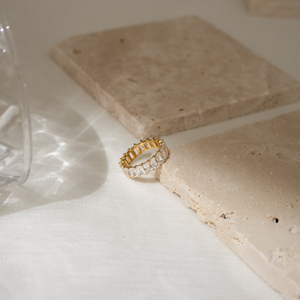 The Colette Eternity Ring, featuring clear baguette diamonds, rests on beige stone tiles in soft natural light, radiating timeless opulence.