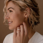 A woman with wavy blonde hair smiles, wearing the Colette Eternity Ring adorned with baguette diamonds and matching stud earrings, radiating elegance against a warm brown background.