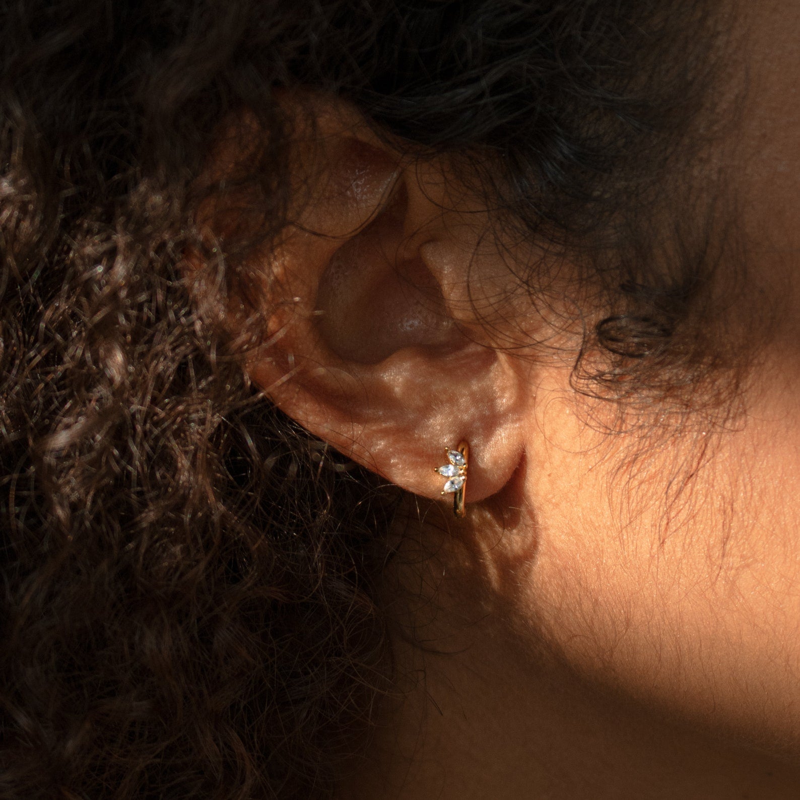 Close-up of a person's ear wearing the Giana Marquise Huggies—small gold hoops with a clear gemstone—shining in sunlight and highlighting the elegance of these subtle earrings.