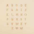 Pave Letter Studs A-Z arranged in rows with a sparkling rhinestone heart and butterfly on a beige background—ideal for creating personalized earrings or adding a touch of minimal style.