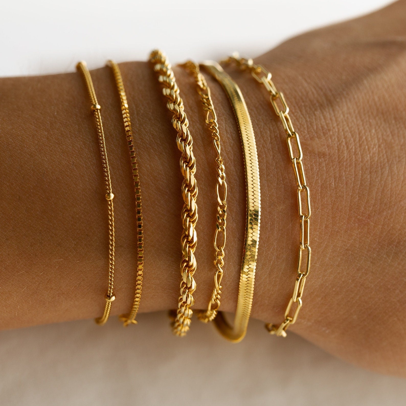 A wrist adorned with six Must-Have Bracelet Chains in various delicate gold styles, including adjustable and trendy paperclip designs.