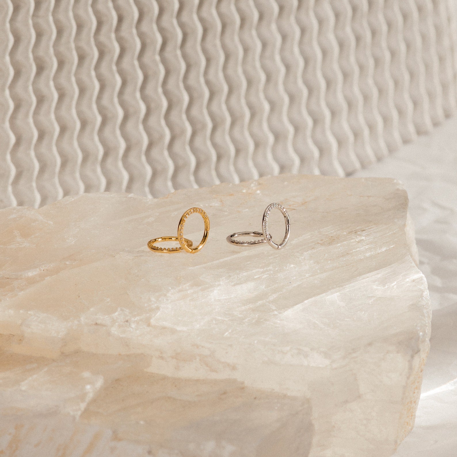 Two minimalist rings—one gold, one silver—are displayed on textured stone with a wavy backdrop, perfectly paired with our Pave Cartilage Mini Huggies for an effortlessly chic look.