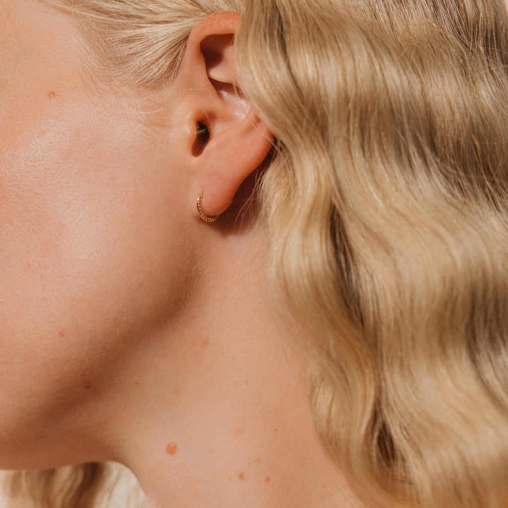 Close-up of a woman's ear wearing Pave Cartilage Mini Huggies, small gold hoop earrings ideal for stacking with other cartilage pieces, paired with wavy blonde hair.
