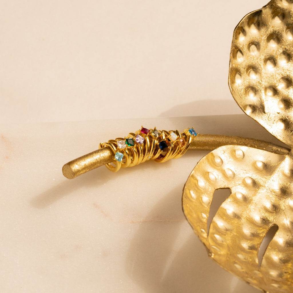 Gold rings with colorful gemstones stacked on a golden branch beside a decorative golden leaf, perfect for pairing with personalized jewelry or Athena Birthstone Huggies.